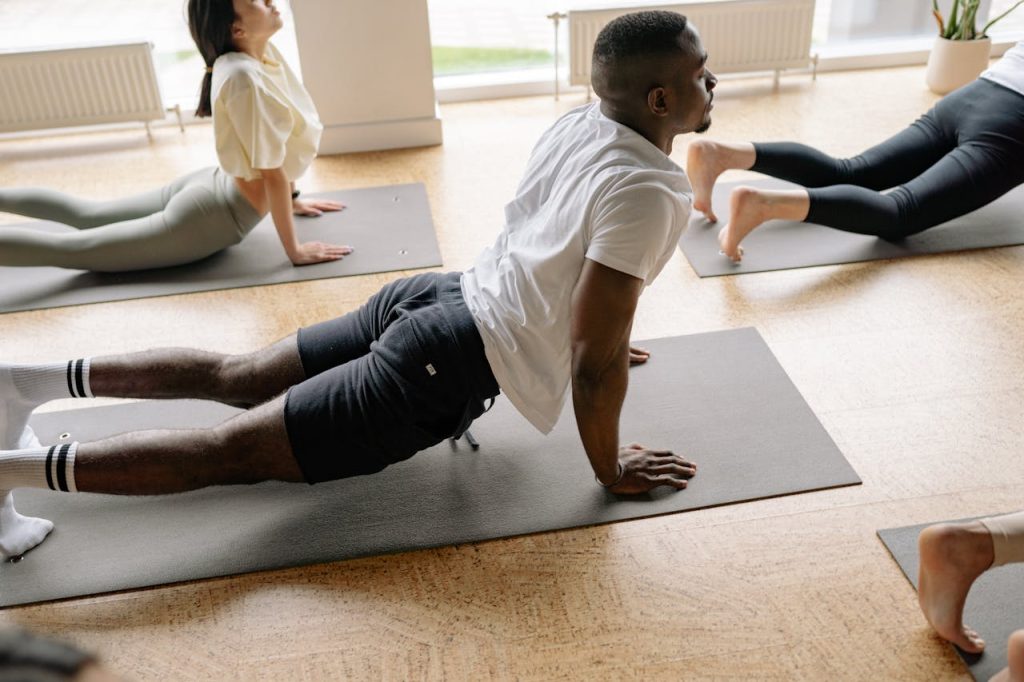 Adults in a yoga class performing stretches on mats in a well-lit studio.